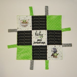 K&ouml;nnte beinhalten: Eine gr&uuml;ne und schwarze Babydecke mit einem karierten Muster. Die Decke hat vier Stoffschlaufen an den Ecken. Die Decke zeigt gestickte Bilder von Figuren aus dem Star Wars-Franchise, darunter Yoda und der Mandalorianer. Der Text "baby Jedi Spencer" ist auf die Decke gestickt.