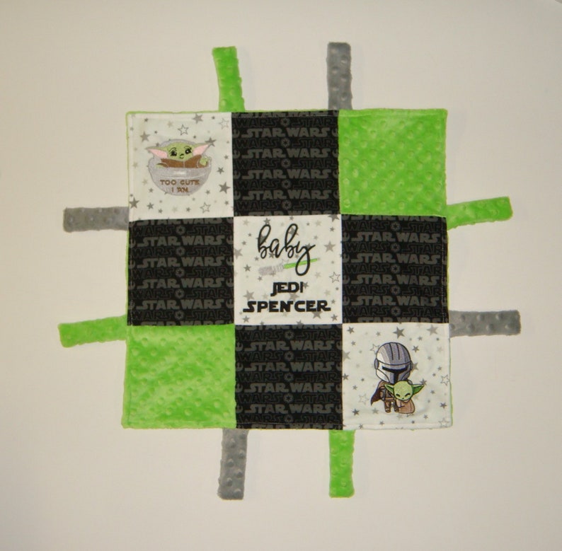 K&ouml;nnte beinhalten: Eine gr&uuml;ne und schwarze Babydecke mit einem karierten Muster. Die Decke hat vier Stoffschlaufen an den Ecken. Die Decke zeigt gestickte Bilder von Figuren aus dem Star Wars-Franchise, darunter Yoda und der Mandalorianer. Der Text "baby Jedi Spencer" ist auf die Decke gestickt.