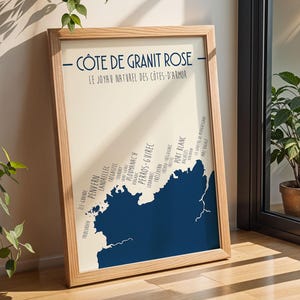Puede incluir: Un póster enmarcado con un mapa de la Côte de Granit Rose en Francia. El mapa es de color azul oscuro sobre un fondo crema, con texto en francés. El marco es de madera clara. El póster está apoyado contra una pared.
