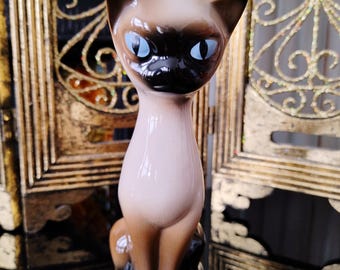 以下が含まれることがあります： 青い目をしたシャム猫のセラミック製置物。猫は尻尾を脚に巻き付けて座っています。置物は茶色と白のグラデーションで塗られています。