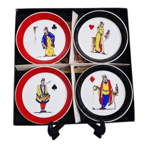 May include: Set of four decorative playing card themed coasters. Each coaster features a different card suit and a whimsical illustration of a royal figure. The coasters have red and black borders and are presented in a black box.