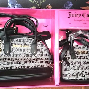 May include: Two Juicy Couture handbags in a pink gift box. The bags are black and white with the brand name printed on them. The larger bag has a gold chain and a black handle. The smaller bag has a black handle.