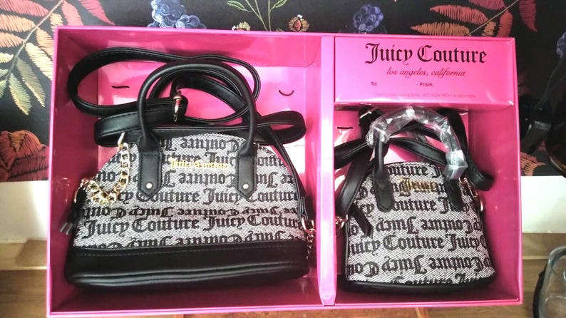 May include: Two Juicy Couture handbags in a pink gift box. The bags are black and white with the brand name printed on them. The larger bag has a gold chain and a black handle. The smaller bag has a black handle.