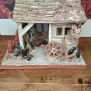 Pode incluir: Figura em miniatura detalhada de um galpão com telhado desgastado e frente aberta, mostrando ferramentas e uma roda de carro. A figura é pintada em tons neutros com detalhes de flores rosa e assenta sobre uma base retangular.