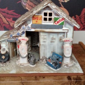 Pode incluir: Modelo de cerâmica detalhado de um posto de gasolina vintage. O edifício em miniatura apresenta um aspeto envelhecido, com letreiros de "Shell" e "Castrol". Inclui duas bombas de gasolina e outros acessórios. O modelo está sobre uma base de madeira.