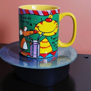 May include: A colorful ceramic mug with a yellow handle and interior. The mug features cartoon illustrations of a cat and a mouse, with text that reads "Buddies Sharing" and "me and my mate who is fab."