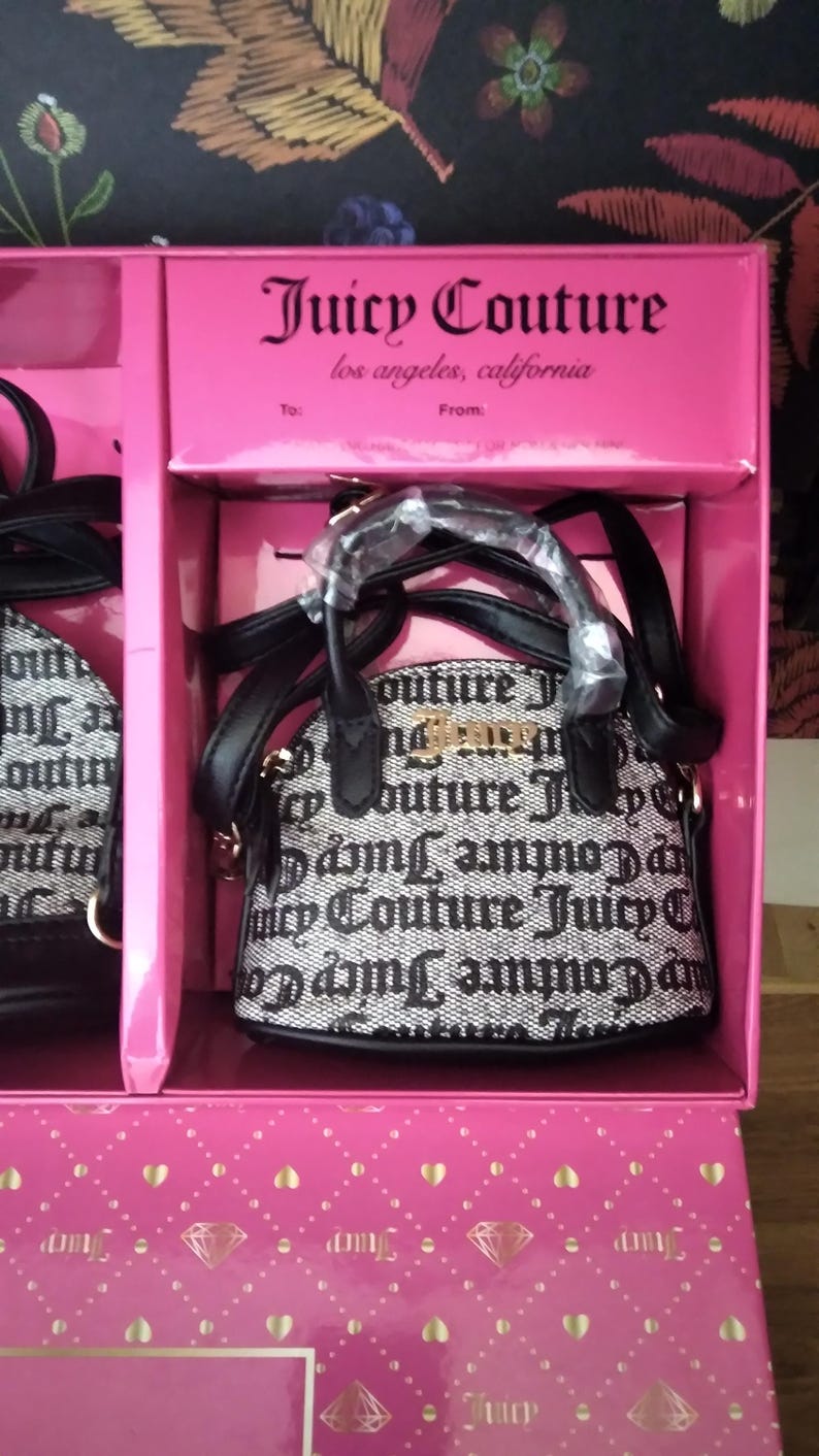 May include: A pink gift box containing a small black and white Juicy Couture handbag. The bag has a repeating logo pattern and gold-colored hardware. The box has the Juicy Couture logo and the text "los angeles, california".