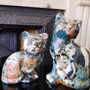 May include: Two ceramic cat figurines, one larger than the other, both covered in a collage of smaller cat images. The larger cat is sitting with its head turned to the side, while the smaller cat is sitting with its head facing forward. Both cats have a white base and are painted in a variety of colors, including black, white, brown, and orange.