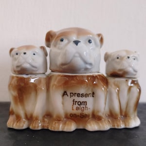 Puede incluir: Un conjunto de tres figuras de bulldog de cerámica. El bulldog del medio es más grande que los otros dos y tiene el texto "A present from Leigh-on-Sea" en su base.