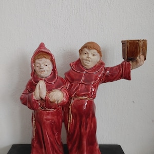Puede incluir: Figura de cerámica que representa a dos monjes con túnicas rojas. Uno está rezando, mientras que el otro sostiene una pequeña caja marrón. Las figuras tienen piel clara y cabello castaño. Las túnicas están detalladas con pliegues y un cinturón de cuerda.