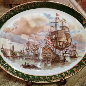 May include: An oval porcelain serving platter featuring a detailed maritime scene of historic sailing ships in a harbor with a city skyline in the background. The platter has a wide, dark green border decorated with an intricate gold filigree pattern.