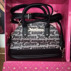 May include: A black and white Juicy Couture handbag with a black handle and strap is displayed inside a pink box. The bag features the Juicy Couture logo in a repeating pattern. The box has a diamond and heart pattern.