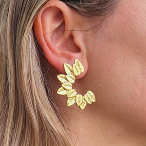 Puede incluir: Pendientes dorados con forma de hojas. Los pendientes están dispuestos en un diseño curvo, con múltiples hojas agrupadas. Los pendientes se llevan en la oreja y la imagen muestra una vista de cerca de la joya.