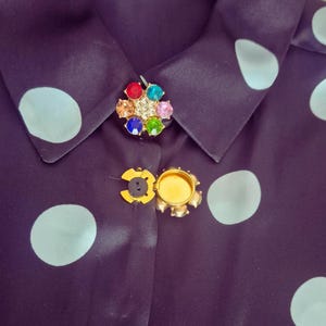 May include: A dark navy blue button-down shirt with large, light blue polka dots. The shirt features two decorative brooch-style buttons, one with colorful gemstones and the other with a gold-colored floral design.