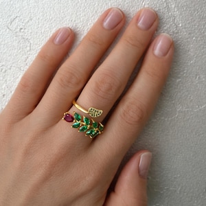 Pode incluir: Um anel dourado com um design de folha com pedras preciosas verdes e vermelhas. O anel envolve o dedo, com um pequeno detalhe de folha na parte superior e um design de folha maior com pedras verdes.