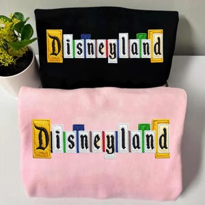 May include: A white ceramic mug with a black handle and the text "But First, Coffee" in a bold, black font. The mug is set against a blurred background. The mug appears to be approximately 10 cm tall.