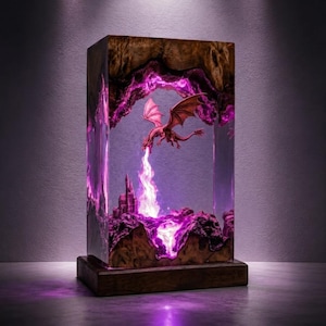 May include: A rectangular lamp featuring a wooden frame with a clear resin center. Inside the resin, a miniature dragon is suspended above a small castle, emitting a bright white light that illuminates the surrounding purple-tinted resin interior.