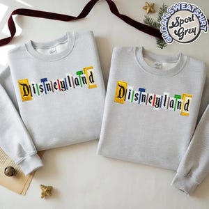 May include: Two light grey sweatshirts with the word "Disneyland" embroidered across the chest in colourful letters. The sweatshirts have long sleeves and a crew neck. The sweatshirts are labelled "Gildan Sport Grey".