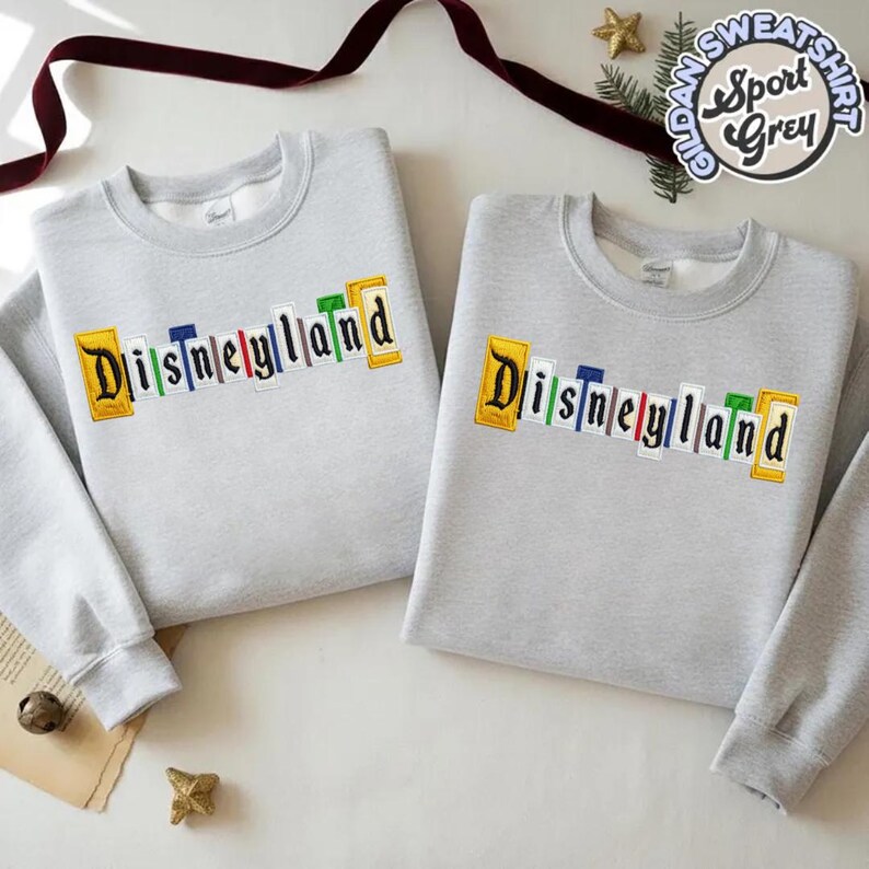 May include: Two light grey sweatshirts with the word "Disneyland" embroidered across the chest in colourful letters. The sweatshirts have long sleeves and a crew neck. The sweatshirts are labelled "Gildan Sport Grey".