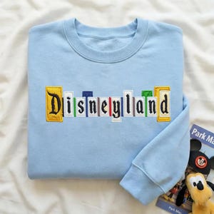 May include: A pale blue sweatshirt featuring the embroidered word "Disneyland" in a colourful, book-style design. It has long sleeves and a crew neck. A small Mickey Mouse toy and a park map are also visible in the image.