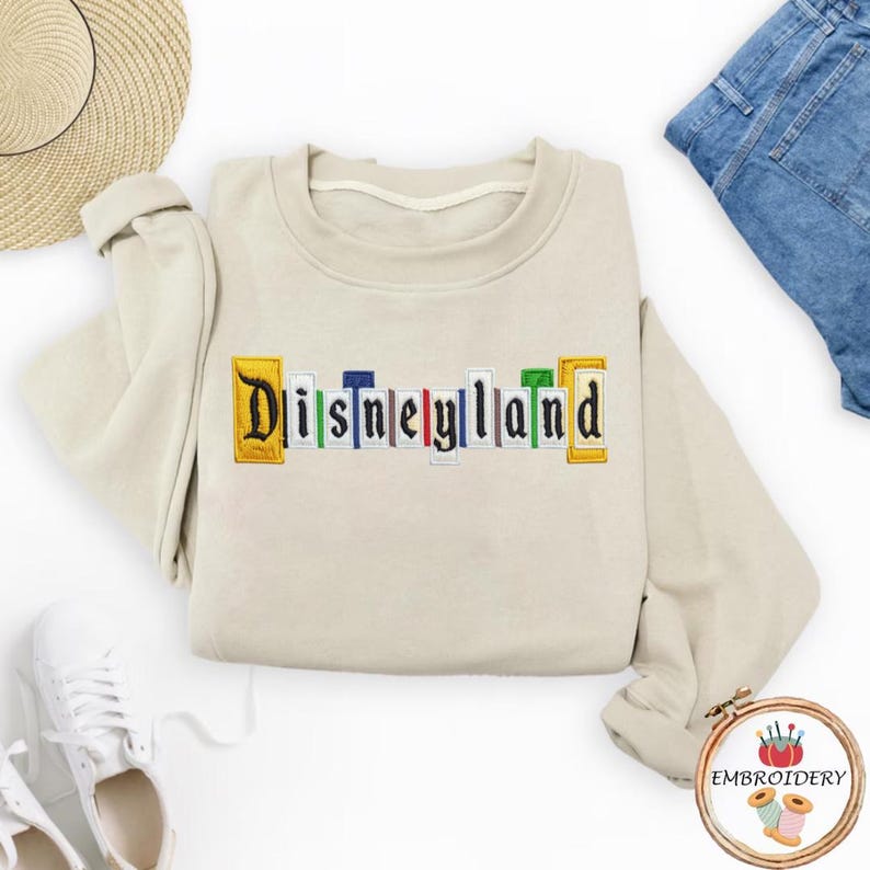 May include: A cream-coloured sweatshirt featuring the embroidered word "Disneyland" in a colourful, block letter design. The letters are outlined in various colours, including yellow, blue, green, and red. The sweatshirt is folded on a white surface.