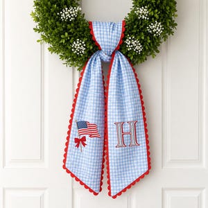 May include: A decorative wreath with a blue and white gingham ribbon featuring a red scalloped edge. The ribbon is embroidered with an American flag and the letter "H". The wreath is made of green foliage and white flowers.