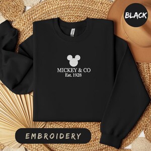 May include: Black sweatshirt with white embroidered Mickey Mouse design and the text "MICKEY & CO Est. 1928". The sweatshirt is displayed on a woven surface with a brown hat in the background. The word "EMBROIDERY" is printed on a black oval.