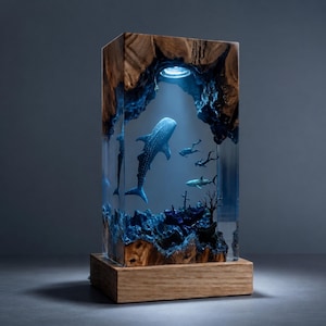 May include: A rectangular lamp featuring a natural wood frame encasing a clear resin block. Inside the resin, a whale shark, two scuba divers, and a smaller shark are suspended above a dark, rocky seabed with a light source at the top.