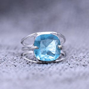 May include: A silver ring featuring a large, square-cut, light blue gemstone. The ring has a split band design, with multiple thin bands creating the ring's structure. The gemstone is the focal point, set in a simple, elegant setting.
