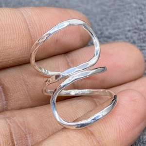 May include: A silver-colored ring with a unique, open design. The ring features an elongated, hammered texture and a spiral shape that wraps around the finger. The ring is shown on a hand.