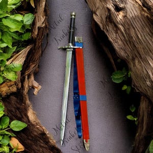 May include: A medieval-style sword with a silver blade, black handle, and a crossguard. The sword is sheathed in a red scabbard with blue accents. The scabbard has a blue strap. The sword is displayed against a dark background with wood and green foliage.