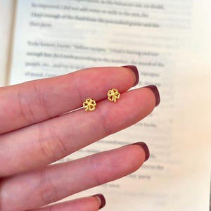 以下が含まれることがあります： 小さな金色のクローバー型のスタッドピアス。ピアスは、手とテキストのページがぼやけた背景に表示されています。クローバーのデザインは、4つの丸みを帯びた花びらを特徴としています。