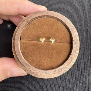 Puede incluir: Un par de pequeños pendientes de botón dorados en forma de corazón, presentados en una caja de joyería redonda de madera con un interior de terciopelo marrón. Los pendientes están centrados en la caja y la veta de la madera es visible en el exterior.