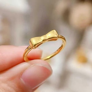 A close-up image of a person's hand holding a gold ring with a bow-shaped charm.