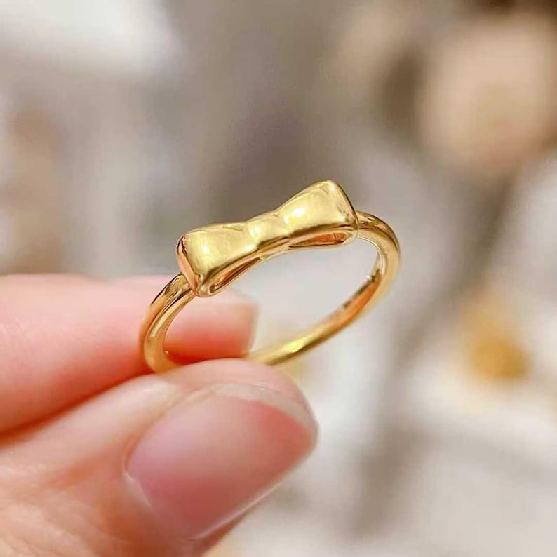 A close-up image of a person's hand holding a gold ring with a bow-shaped charm.