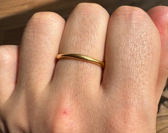May include: A thin, smooth, gold-colored band ring with a subtle, gentle wave or curved silhouette. The ring features a polished, reflective surface that catches the light, creating a simple and continuous circular form around the finger.