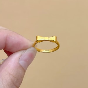 A gold ring with a bow-shaped design is being held in a person's hand against a beige background.