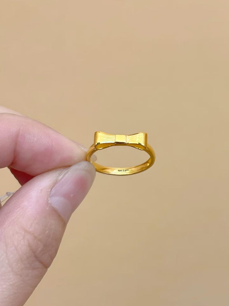 A gold ring with a bow-shaped design is being held in a person's hand against a beige background.