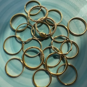 Puede incluir: Una colección de anillos dorados con diversos diseños, incluyendo bandas lisas, trenzadas y con texturas. Los anillos están dispuestos sobre una superficie azul claro, creando una exhibición visualmente atractiva de joyería.