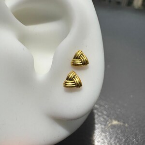 May include: A pair of gold-coloured stud earrings, each featuring a small, triangular shape with a textured, woven-like pattern. The earrings are displayed on a white ear-shaped model, showcasing the metallic finish and the intricate, knotted design.