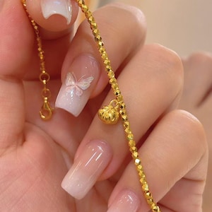 Può includere: Un delicato braccialetto d'oro con un design di perline sfaccettate e un piccolo ciondolo. Il braccialetto è tenuto in alto, mostrando i suoi dettagli intricati e la calda tonalità dorata del metallo. Il ciondolo ha una piccola forma rotonda.