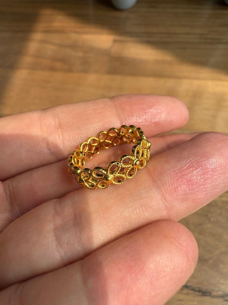A gold ring with a delicate, intricate design is being held in a person's hand against a wooden background.