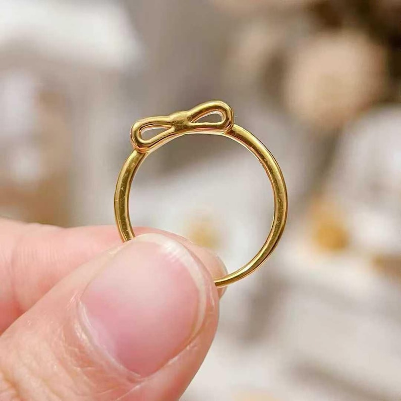 A close-up image of a person's hand holding a gold ring with a bow-shaped design on the top.