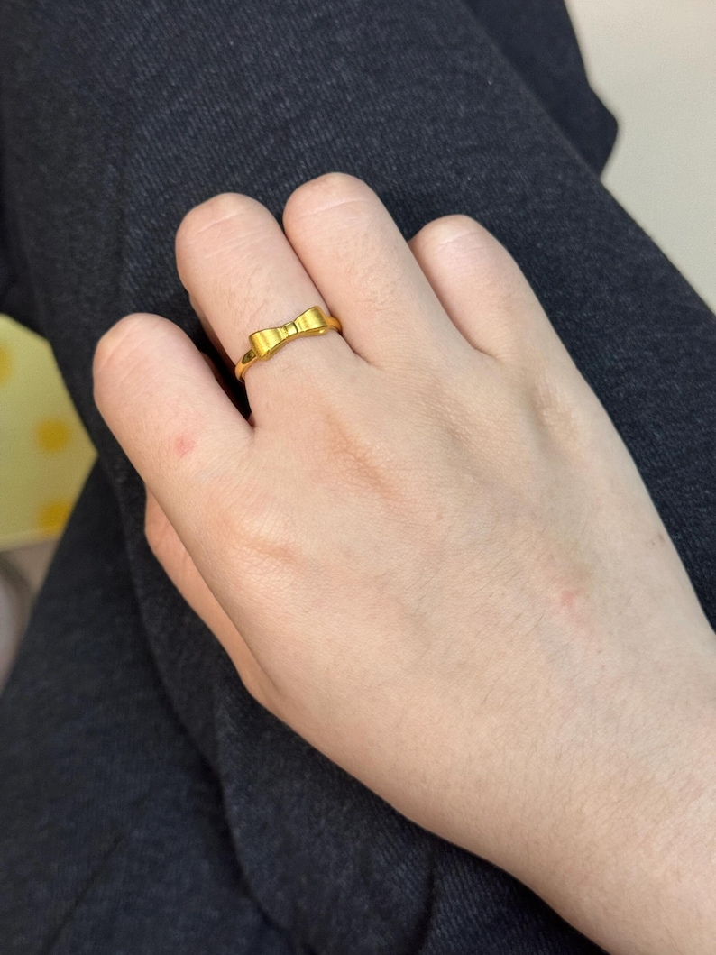 A close-up view of a person's hand wearing a gold ring with a bow design.