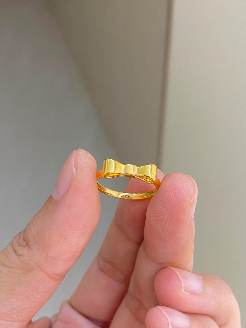 A person's hand holding a gold ring with a bow design.