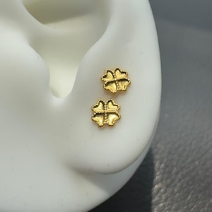May include: A pair of gold-toned stud earrings shaped like four-leaf clovers. Each earring features a textured, beaded border around the edge of the clover petals and a smooth, recessed center, creating a distinct metallic finish on the surface.