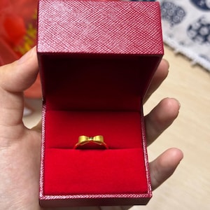 A hand holding a red jewelry box containing a gold ring.