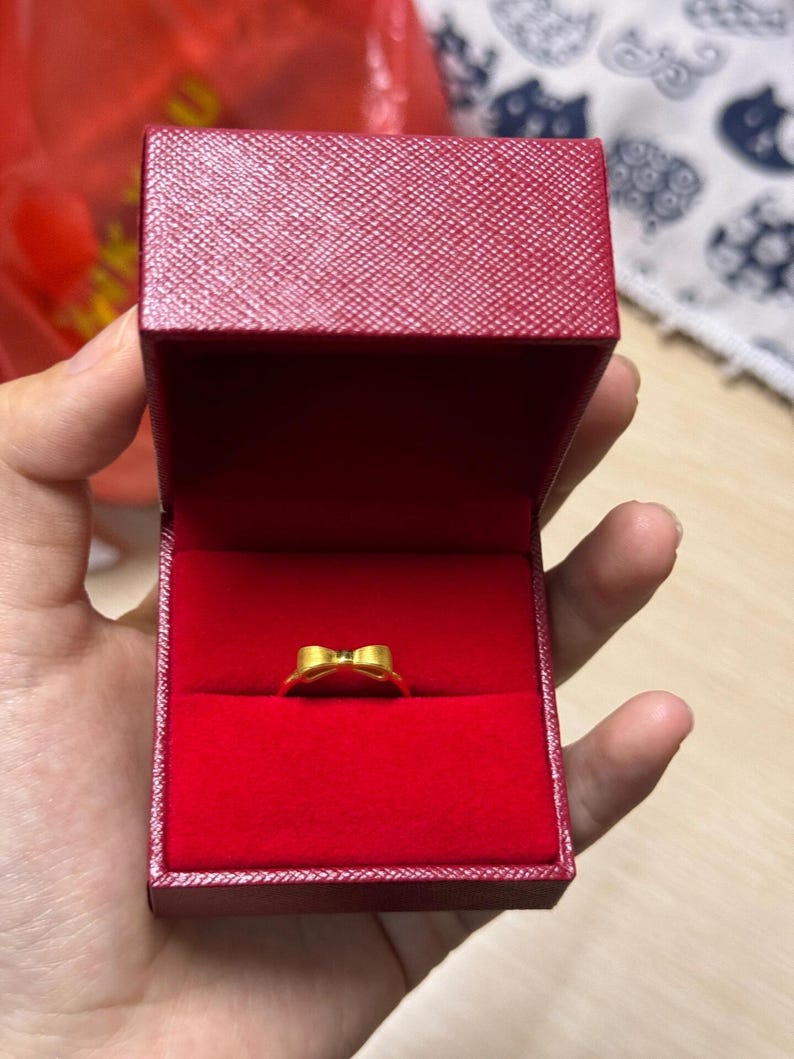 A hand holding a red jewelry box containing a gold ring.