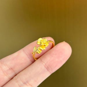 A gold ring with a flower design is being held in a person's hand against a blurred green background.