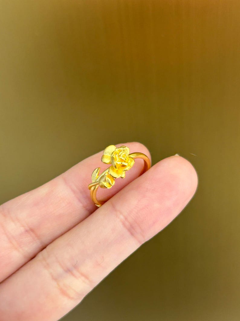 A gold ring with a flower design is being held in a person's hand against a blurred green background.